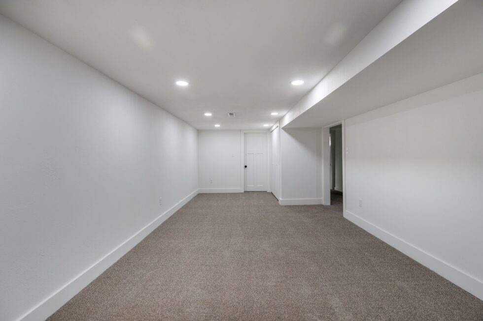 A renovated, empty, and clean basement with white walls and fresh carpet, showing the result of a successful audit and cleanout.