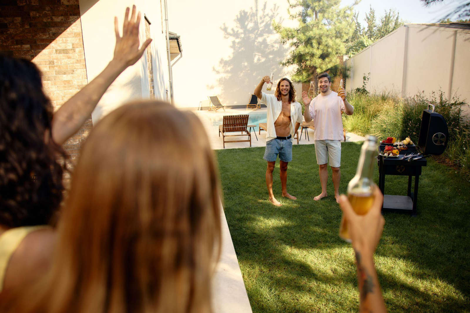 A group of friends enjoying a backyard barbecue and raising their drinks on a clean, green lawn next to a swimming pool.