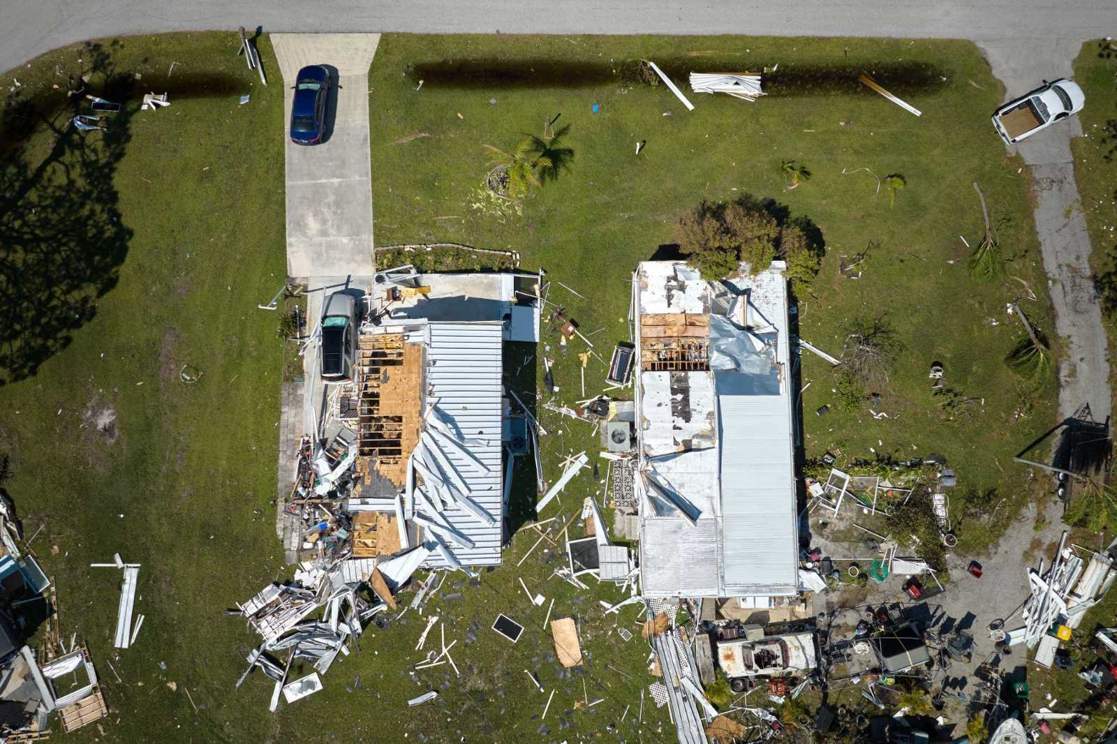 Top-down drone view of houses with severe roof damage and debris scattered across yards following a severe storm.