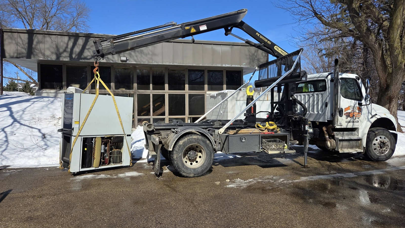 A white Junk Truck equipped with a boom crane lifting a heavy commercial appliance outdoors during a winter cleanup.